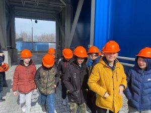 Grupa chłopców w pomarańczowych kaskach maszeruje na tle niebieskiego budynku