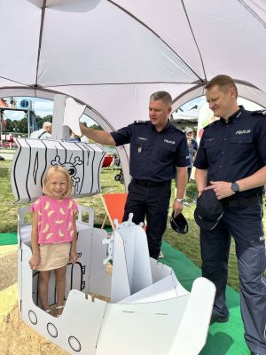 Dziewczynka w różowej bluzce stoi wraz z dwoma policjantami w granatowych mundurach przy białym, kartonowym statku