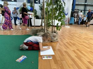 Dziewczynka w czerwonych spodniach klęczy na zielonym dywanie i rysuje kredkami na kartce